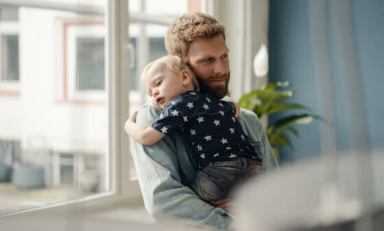 Father standing in front of a window holds his child as it sleeps on his shoulder