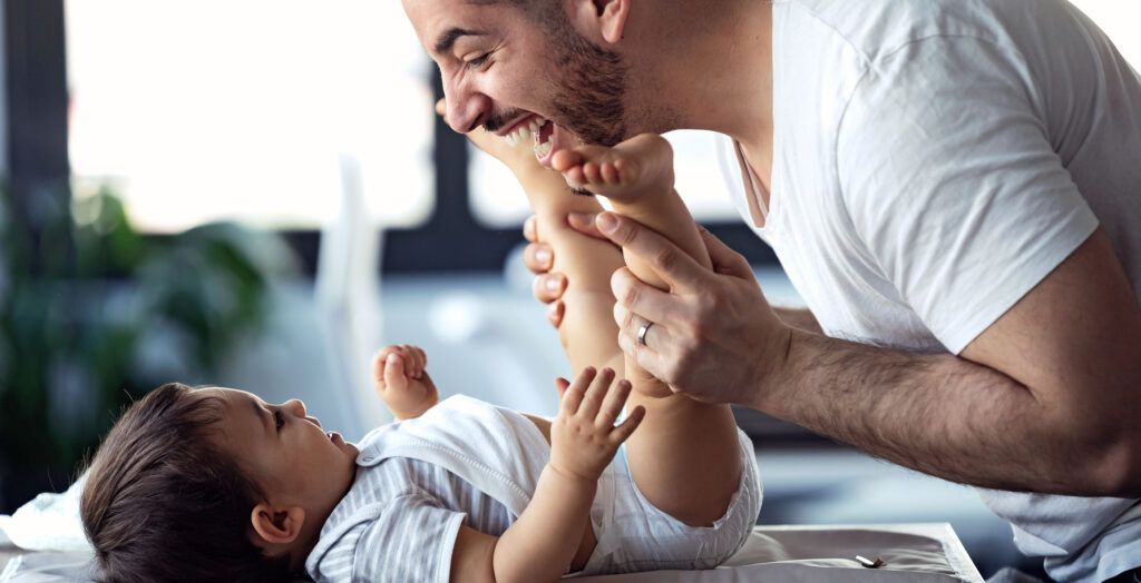 Dad laughing with and grabbing baby’s legs after a nappy change.