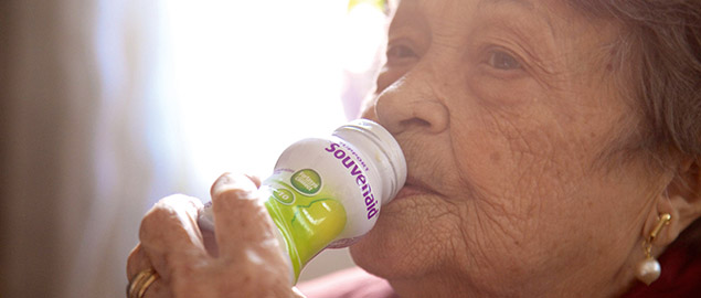 An elderly woman drinks from a bottle of Souvenaid Liquid.