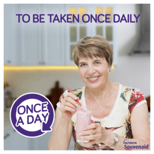 A smiling woman holds a glass of Souvenaid in her kitchen. A ‘Once a day” logo is in the corner.